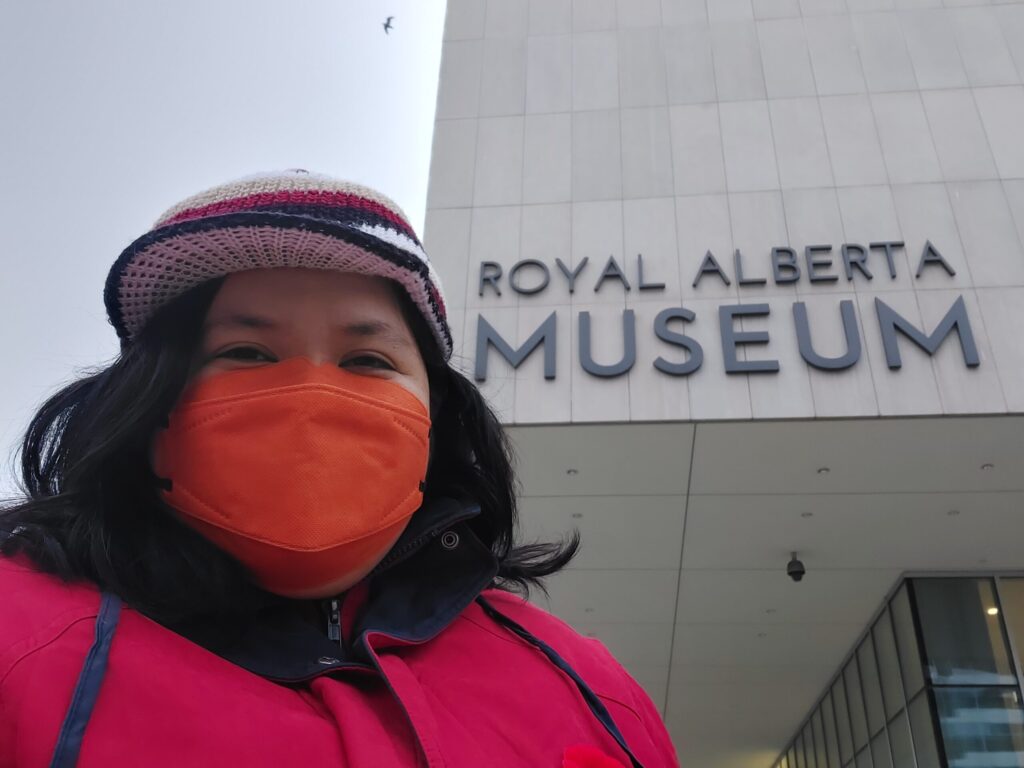 Giselle General, a Filipino woman with shoulder length dark hair wearing a knit cap, pink coat, and orange face mask, standing outside the Royal Alberta Museum. Museum sign is visible in background.