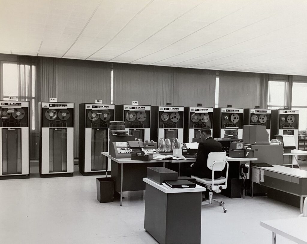 A room with nine tall computer cabinets along the wall. A man wearing a suit sits at a worktable in front of the computer. The table holds large computer tapes and other equipment needed to run the 7070.