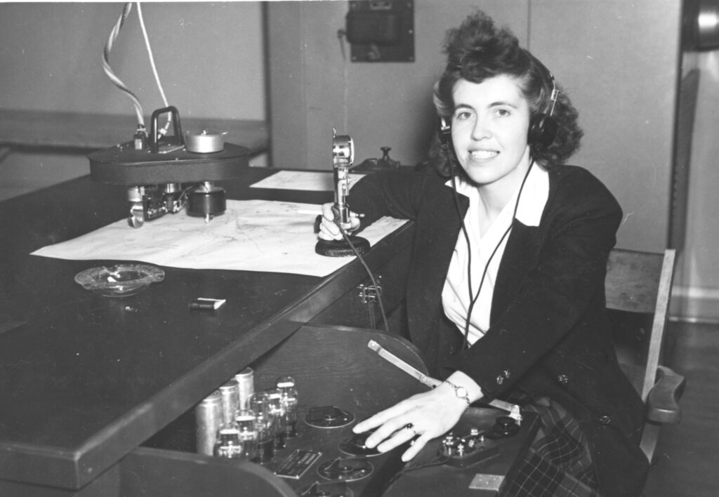 Margaret Littlewood, a young white woman with dark, shoulder-length hair, sits at the Link trainer desk. The angle of the photo emphasizes the technical aspects of the machine. Littlewood’s right hand is grasping what appears to be a kind of microphone that is sitting on the desk. She is wearing a white collared shirt, dark sweater, and plaid skirt.