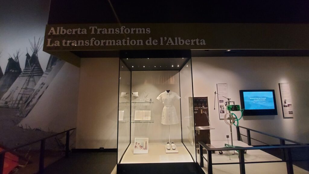 A photograph of a Royal Alberta Museum exhibit called “Alberta Tranforms/La transformation de l’Alberta. In the center of the photograph is the exhibit featuring the work of Tessie Oliva.