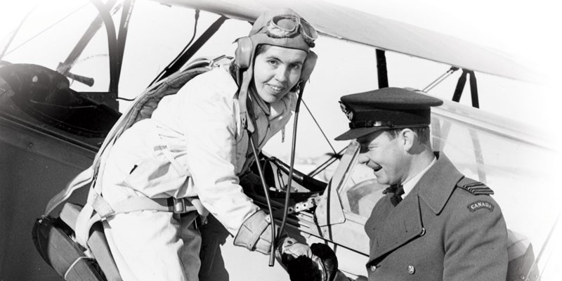 Margaret Littlewood, wearing aviation gear, steps out of a small prop plane. She is looking at the camera and grasping the hand of an air force officer dressed in a military coat and hat and wearing thick gloves.