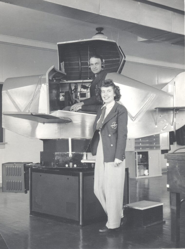 Margaret Littlewood, a young white woman with dark hair, stands in front of the Link trainer, which is a flight simulator machine. She is wearing a uniform that include a tie and jacket. A white man sits in the airplane portion of the trainer behind Littlewood, wearing headphones, looking at the camera.
