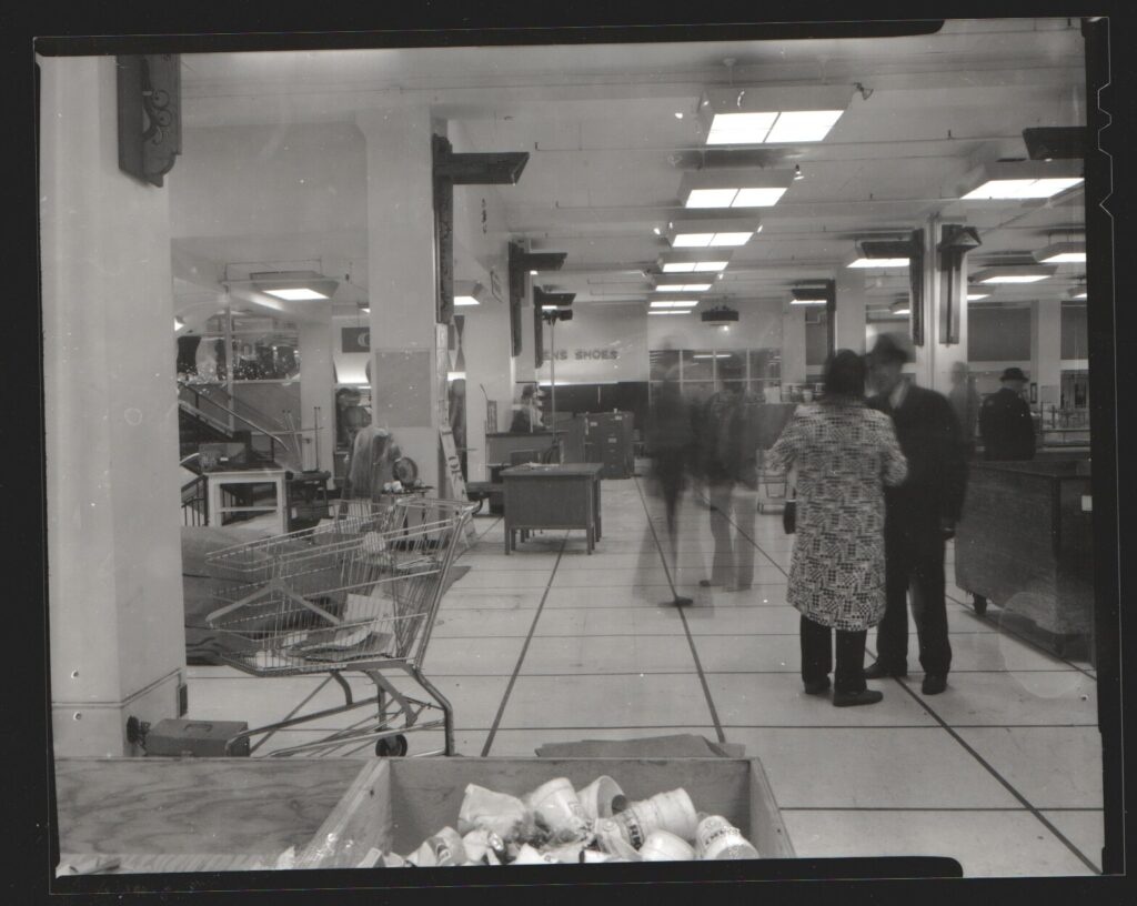 An internal scene from 1974 in Woodward’s department store in Edmonton. The photograph shows customers, a shopping cart, and merchandise.