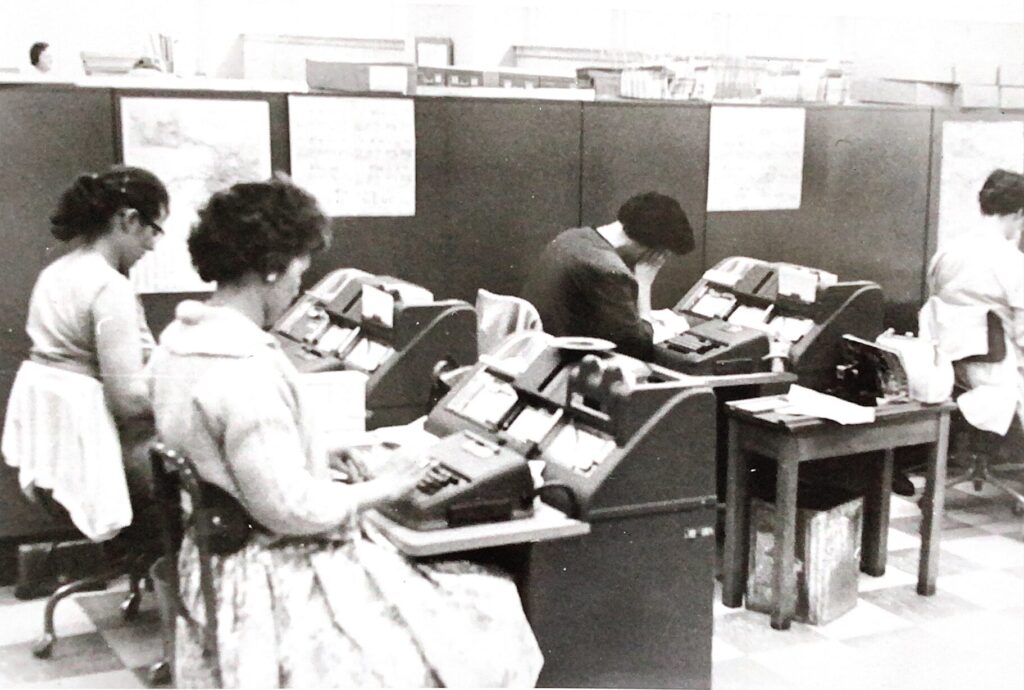 Four women sit at workstations in an office setting entering data on key punch machines. One worker, bent forward, appears to be resting with her head in her hand.