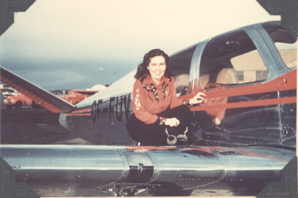 Margaret Littlewood, a white woman with long dark hair, at the side of a small silver plane with red accents and “CF-FUV” printed on the side of the aircraft. Littlewood is dressed in a long-sleeved red shirt that matches the plane and black pants.