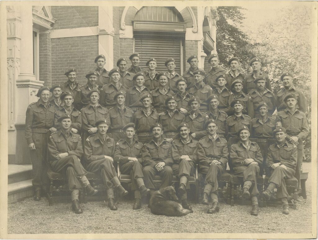 A large group of military officers, members of the Loyal Edmonton Regiment, pose in uniform for a photo. There is a dog laying on the ground in the front of the group.