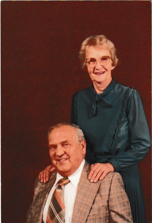 A white couple in their sixties posed together. The man is sitting, wearing a tan suit, white collared shirt, and plaid tie. The woman is standing angled behind him with her hands on his shoulders. Her hair is short and blond; she is wearing glasses and a long-sleeved teal dress.