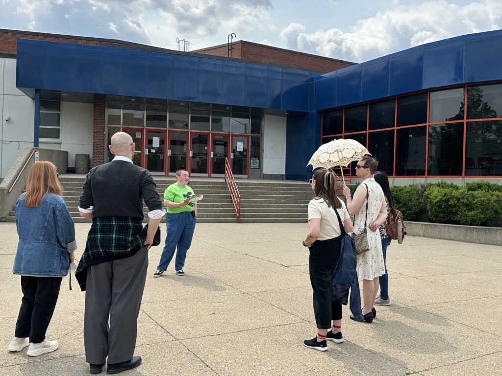 A photograph of a small group of people standing in front of Victoria School for the Arts. Abigail addresses the group from the middle.