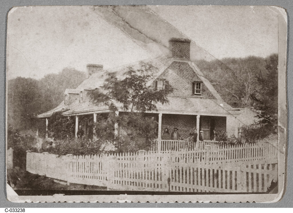 Daguerreotype of a woman and her two children on the verandah of a two-story house, with trees and white fences.