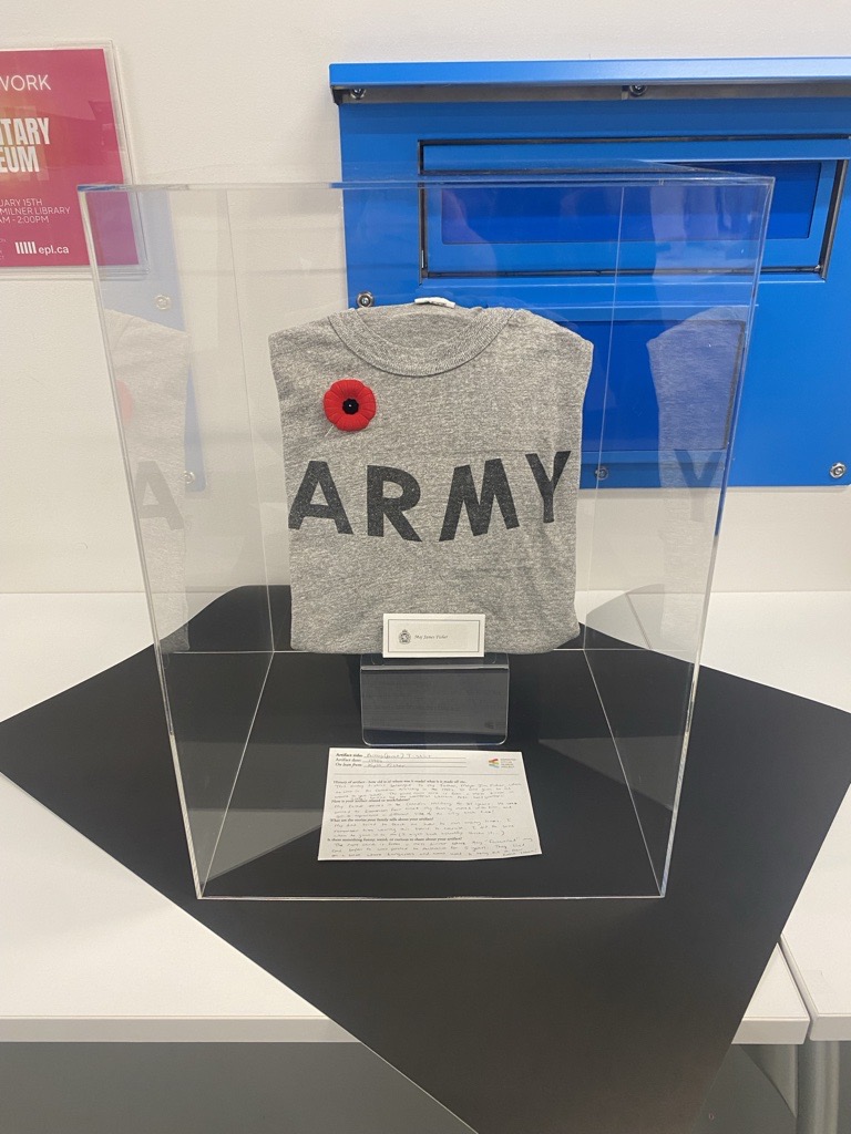 A photo of a grey t-shirt that says ARMY, with a poppy pinned on, in a display case.