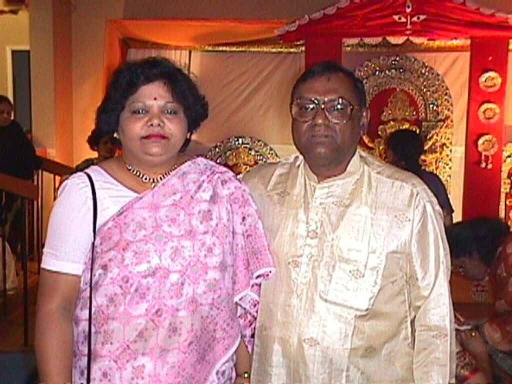 Sanjukta and Dilip, a middle-aged South Asian couple, in formal party wear posing in front of a decorated backdrop.