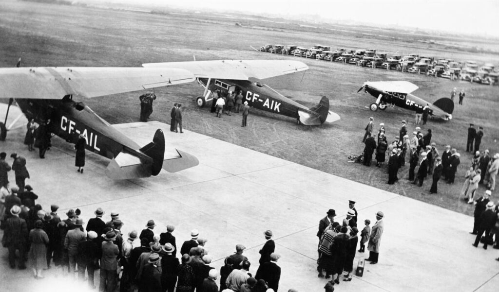 The Grads prepare for departure. Crowds stand on the Blatchford Airfield near three small airplanes. A row of cars is parked in the background.