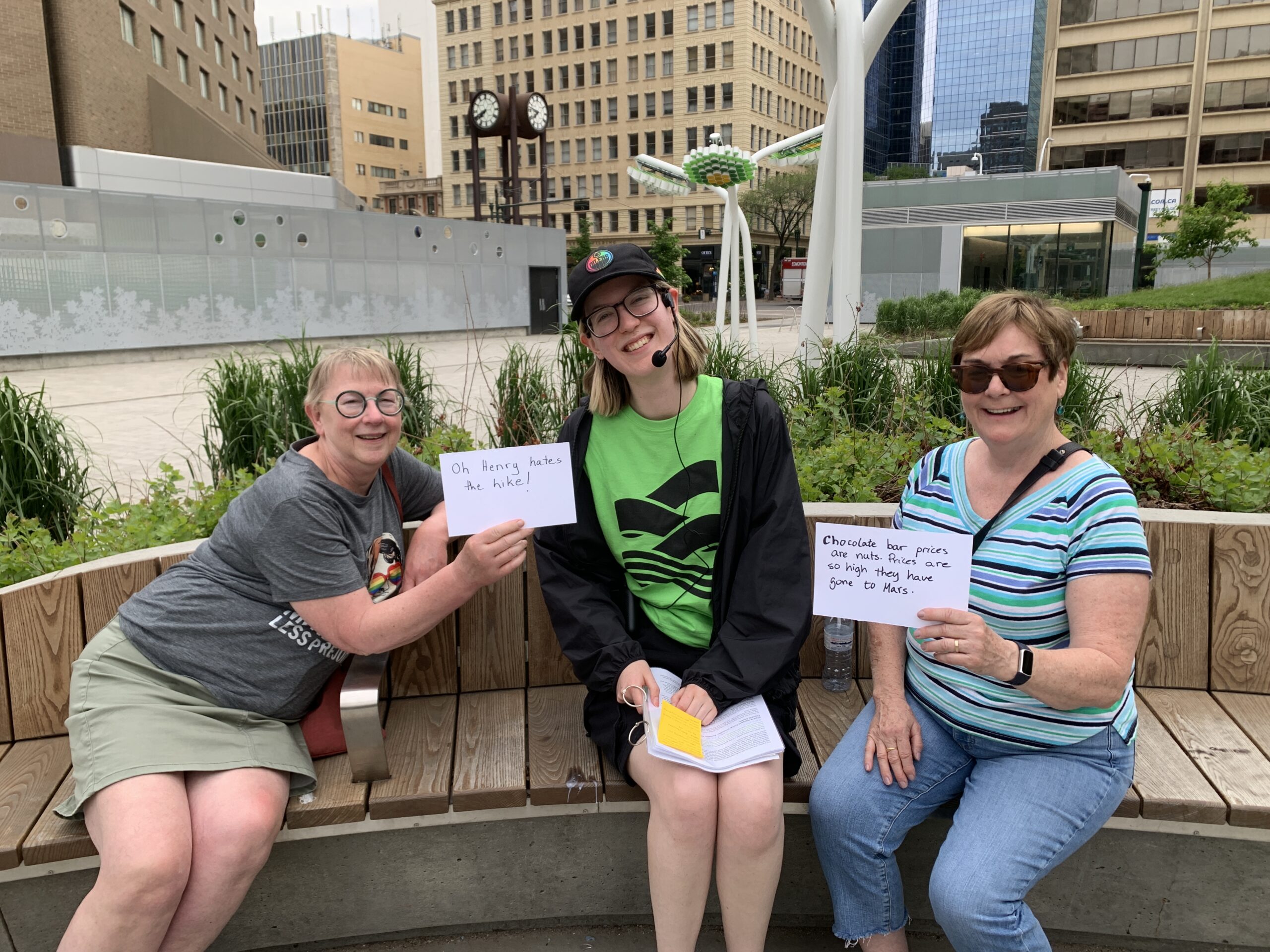 A photograph of three people smiling on a bench in Centennial Plaza. Emily smiles in the middle, in a green ECAMP tshirt. Two smiling tour attendees hold up protest signs about chocolate bar prices.