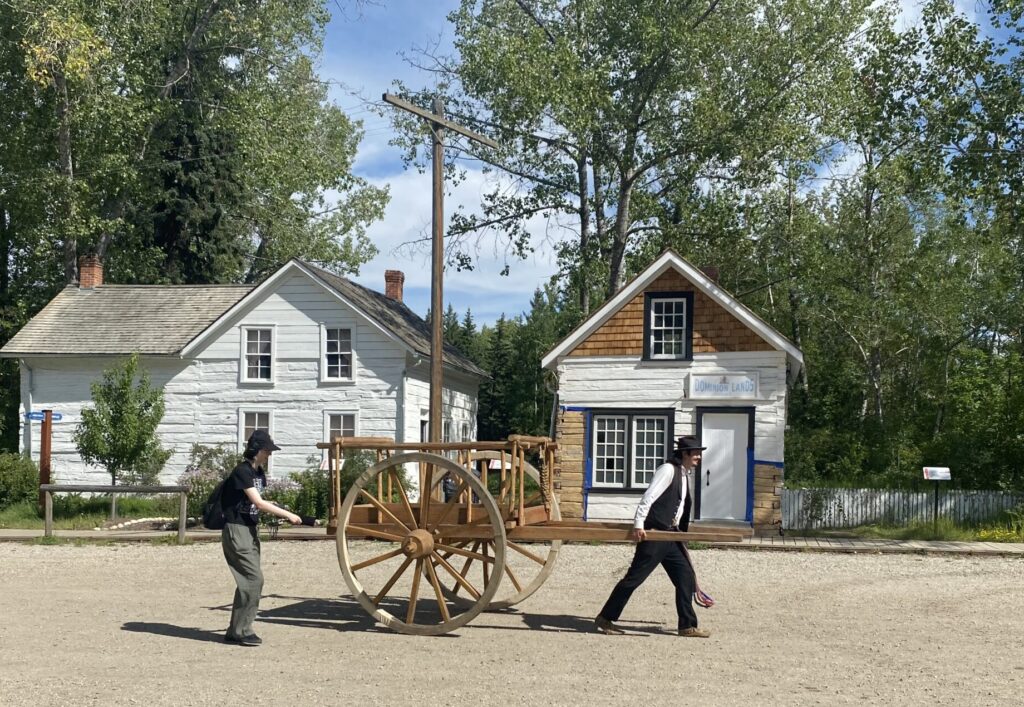 Emily, wearing headphones and holding out a microphone, follows an interpreter in traditional Metis attire who’s pulling a large-wheeled, wooden cart.