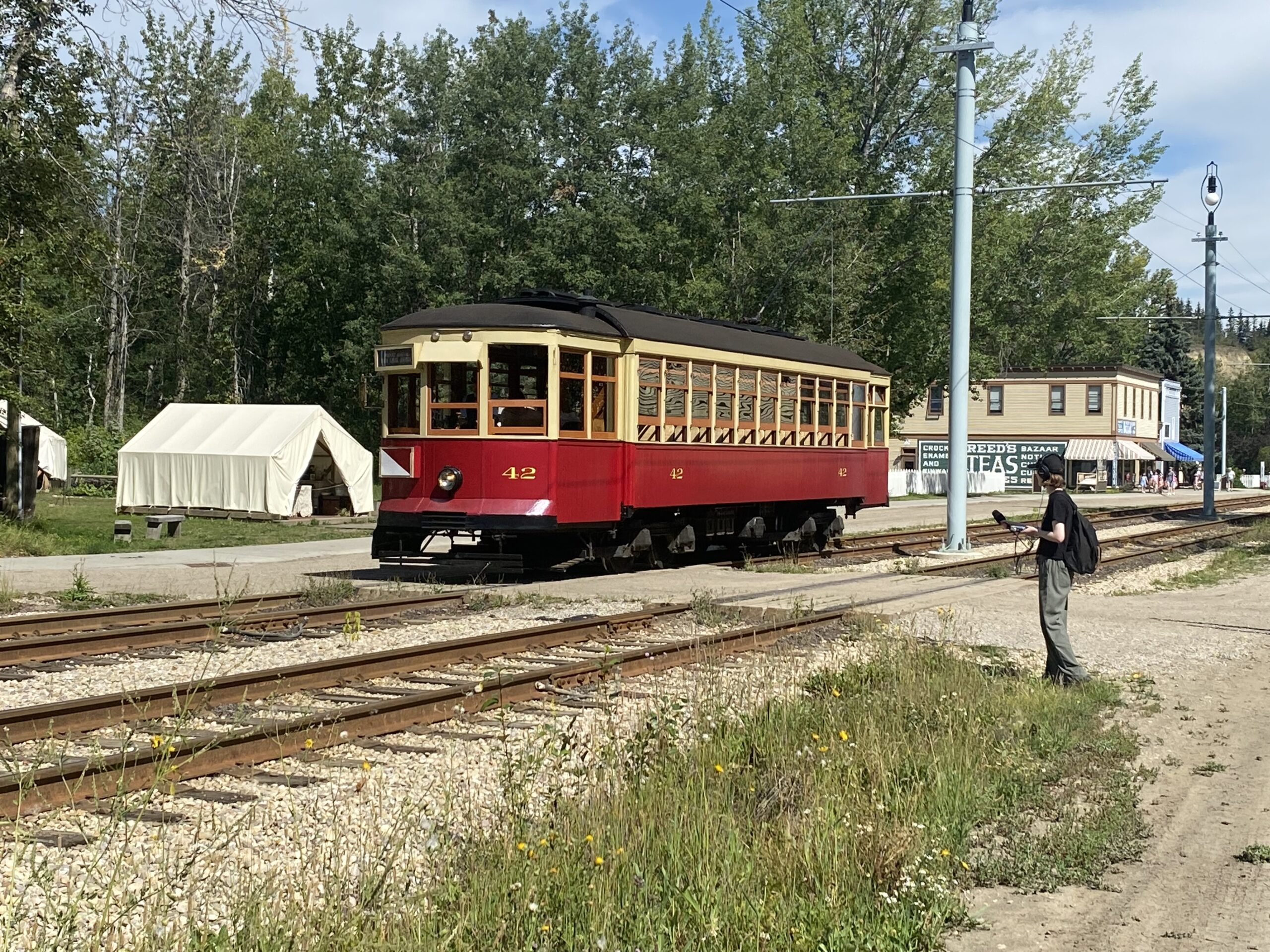Emily holds a microphone out towards a white-and-red streetcar that’s passing them on a railway track.
