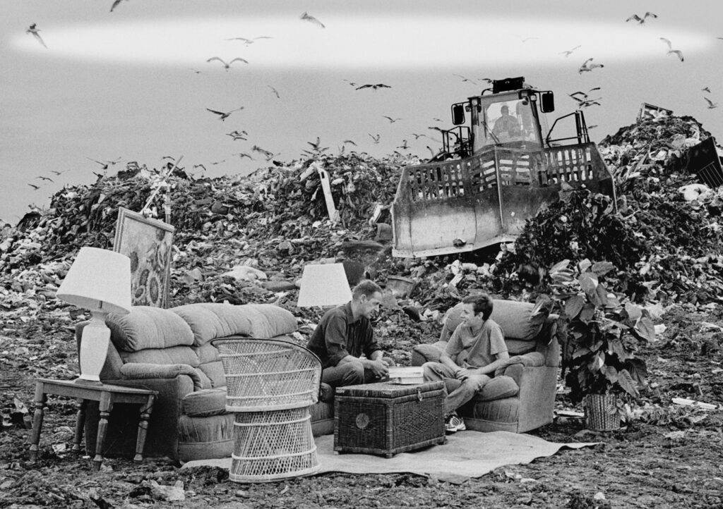 :  A candid photo of a middle-aged man and teenage boy sitting opposite each other on living room furniture in the middle of a landfill. There is a trash compactor in the background, as well as an immense pile of garbage.