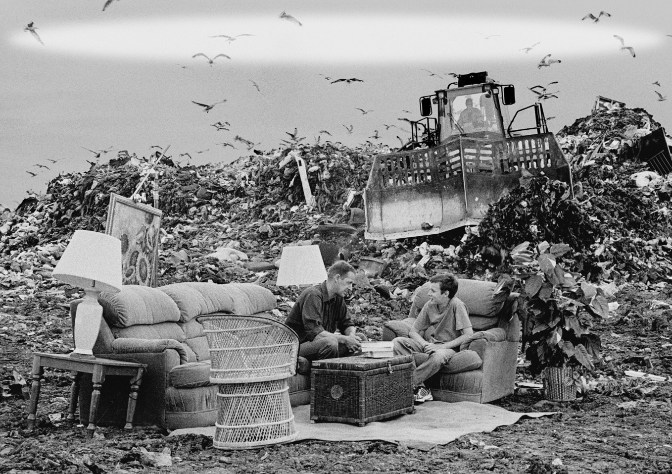 : A candid photo of a middle-aged man and teenage boy sitting opposite each other on living room furniture in the middle of a landfill. There is a trash compactor in the background, as well as an immense pile of garbage.