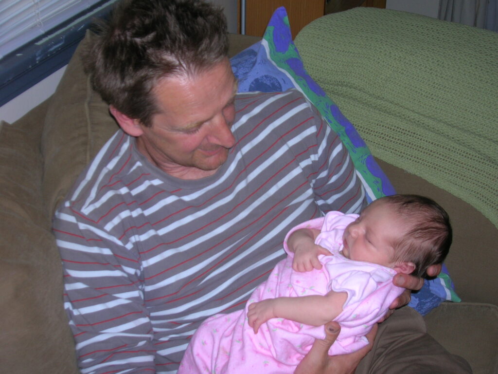 A candid photo of the same middle-aged man as the last photo, sitting on a brown couch and holding a newborn baby. He’s looking down at the baby in his arms, who’s asleep.