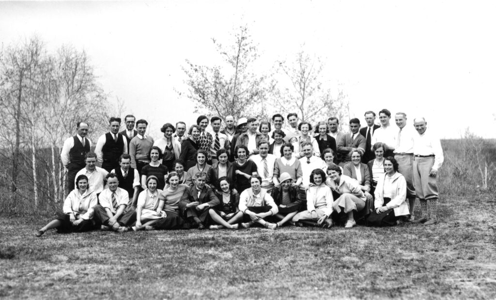 A photograph of a large group of men and women posing on the grass in front of some trees.