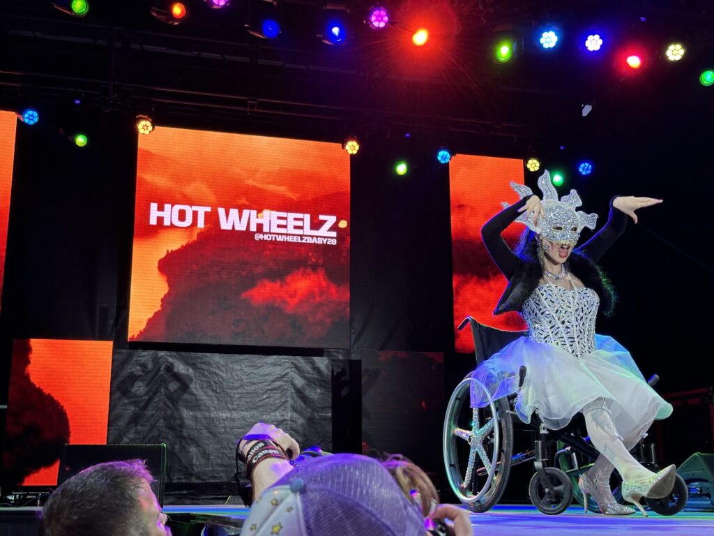 Hot Wheelz lipsyncs in a jewel-encrusted, pointed head dress, corset, skirt, and high heels. They are in a wheelchair on stage above a crowd taking pictures.
