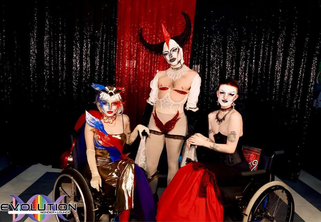 Three drag performers in arch outfits and makeup. Red Silence stands in the centre with horns and nipples exposed. Hot Wheelz and Aboyactually pose in wheelchairs on either side in lowcut dresses.
