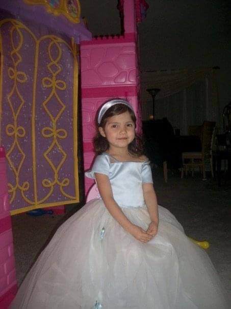 A small child with a feminine appearance poses and smiles in a conventional light blue dress smiles at the camera.