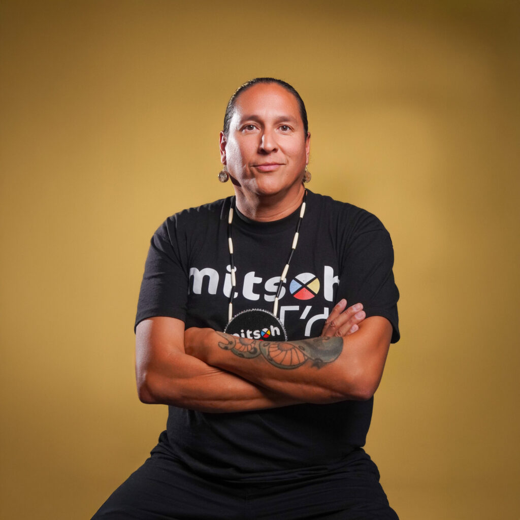 A man in a Mitsoh t-shirt and necklace poses with his arms crossed.