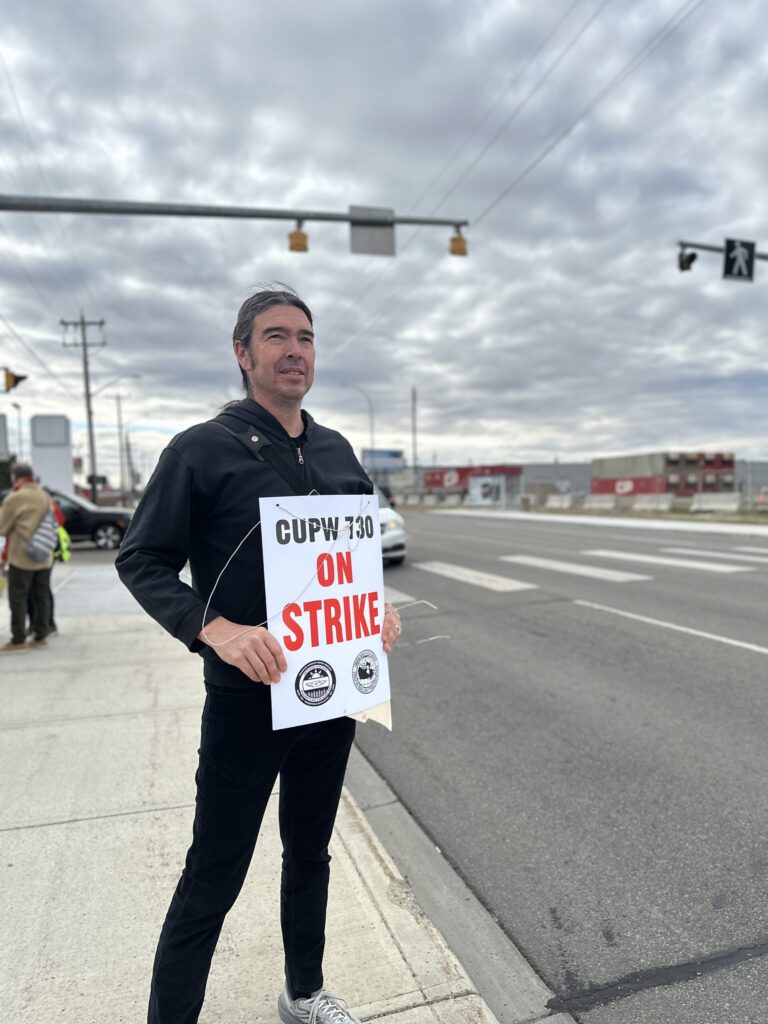A man stands on the sidewalk as traffic goes by. He is holding a sign that says CUPW 730 – On Strike.