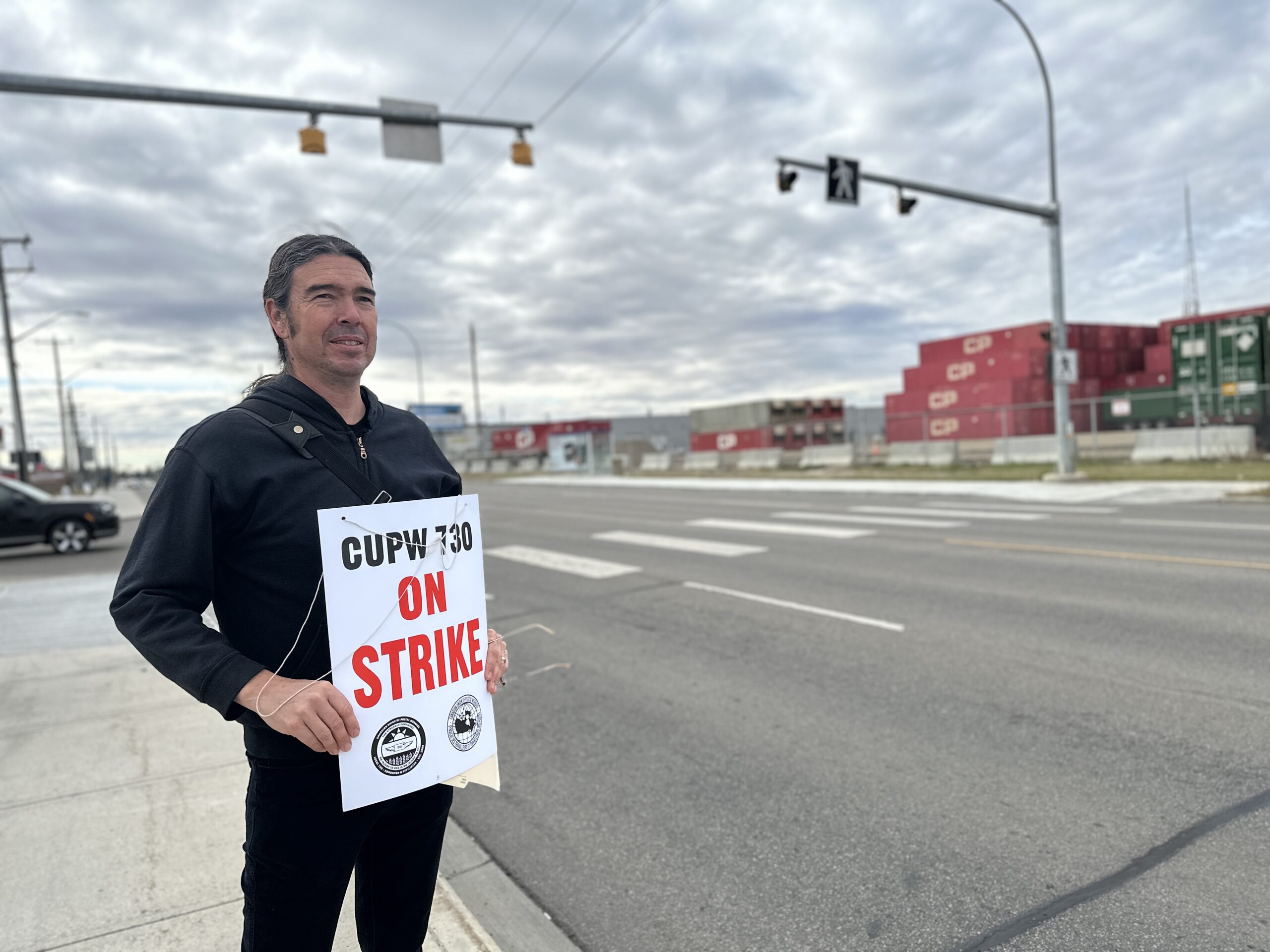 A man stands on the sidewalk as traffic goes by. He is holding a sign that says CUPW 730 – On Strike.