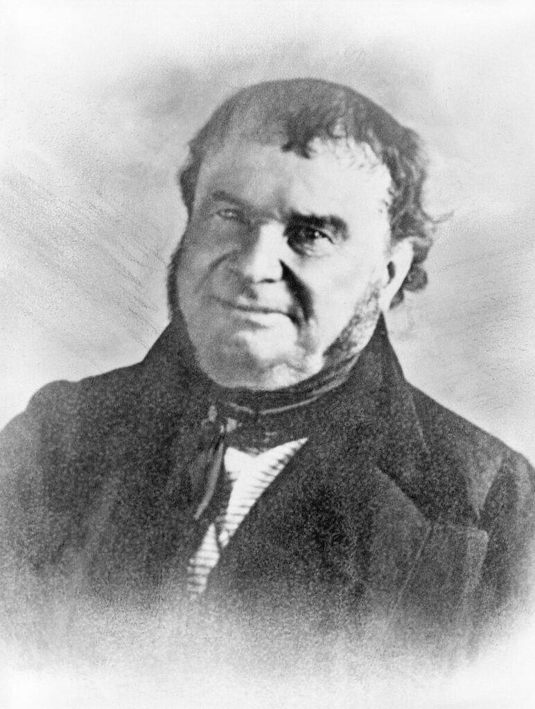 Photograph. A smiling broad-faced man with mutton chops poses for a photograph in a black suit.