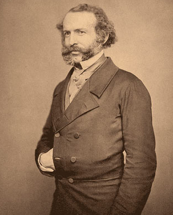 A white man with mutton chops and a moustache poses for a photograph with one hand inside his double-breasted coast.