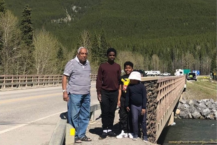 Four people posing for a photo on the side of a two lane paved bridge on a sunny day, with heavily forested mountains behind them.