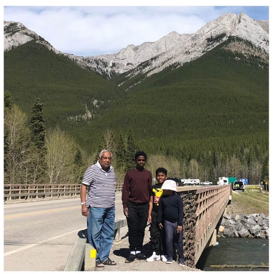 Four people posing for a photo on the side of a two lane paved bridge on a sunny day, with heavily forested mountains behind them.