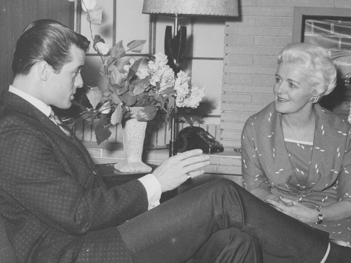 A photograph of Robert Goulet in a suit with slicked-back hair speaking to Laura Lindsay, who is elegantly dressed in front of a fireplace.