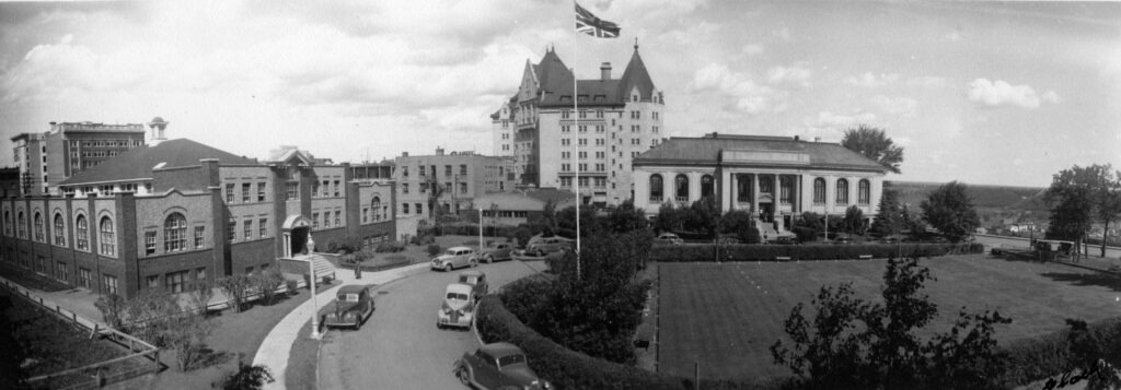 A black and white panoramic photograph of Edmonton in 1924. Flanking a public green space in the foreground are two stone buildings. A simple brick building for veterans on the left-hand side and a neo-classical style library in the mid-ground. Tall in the background is the châteauesque-styled Hotel MacDonald. 