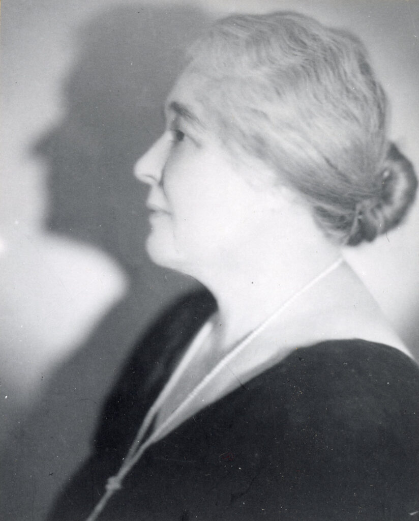 A black and white photograph of Maud Bowman - a middle-aged woman shown in profile from the shoulders up. She wears a v-cut black top with a long white pearl necklace and her hair is pulled back into a low bun.