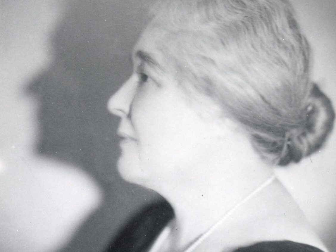 A black and white photograph of a middle-aged woman shown in profile from the shoulders up. She wears a v-cut black top with a long white pearl necklace and her hair is pulled back into a low bun.