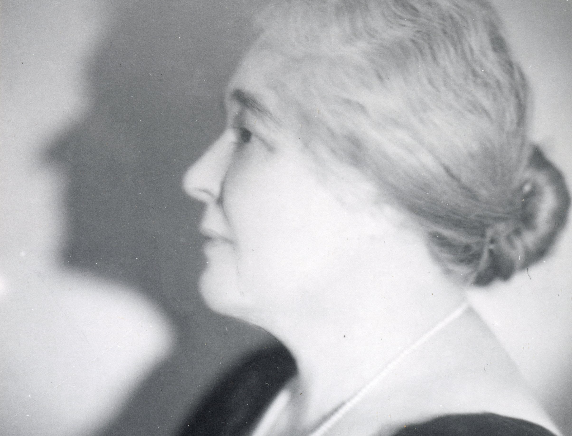 A black and white photograph of a middle-aged woman shown in profile from the shoulders up. She wears a v-cut black top with a long white pearl necklace and her hair is pulled back into a low bun.