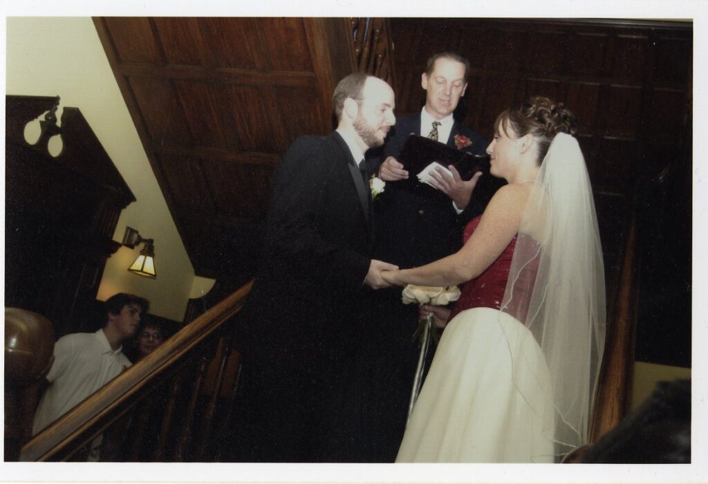 Three people are standing on a staircase surrounded by ornate wood paneling. One is the groom, in a black suit with a white rose boutonniere, and the other is the bride, in a wedding dress with a red, beaded bodice and white veil, holding a bouquet of white roses. The officiant stands between them in a suit, reading from a black portfolio.