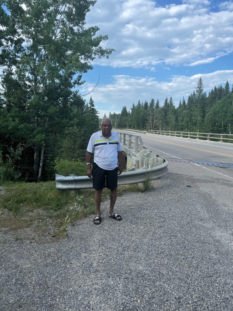 An older man standing in flip flops by a two lane paved bridge on a sunny day.