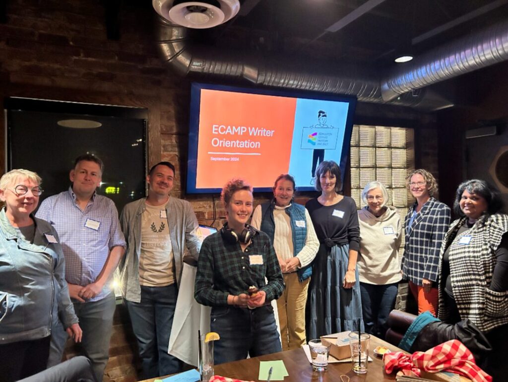 Writers pose in a line in a brick room, in front of a TV screen with a slide that says ECAMP Writer Orientation