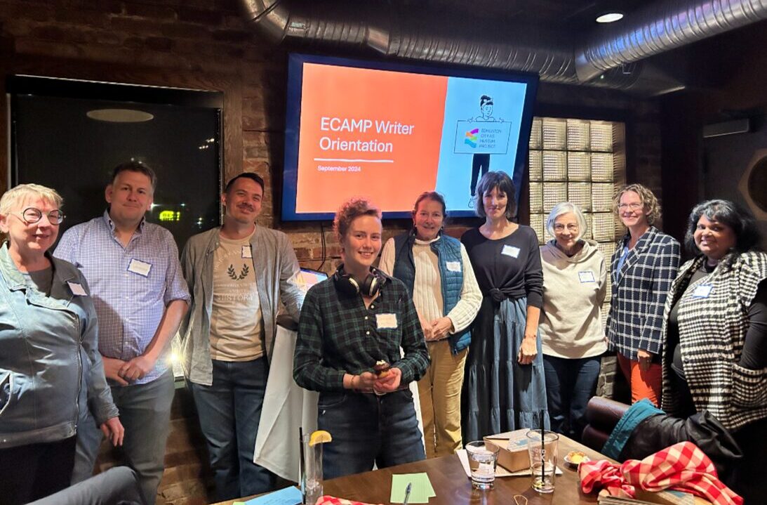 Writers pose in a line in a brick room, in front of a TV screen with a slide that says ECAMP Writer Orientation