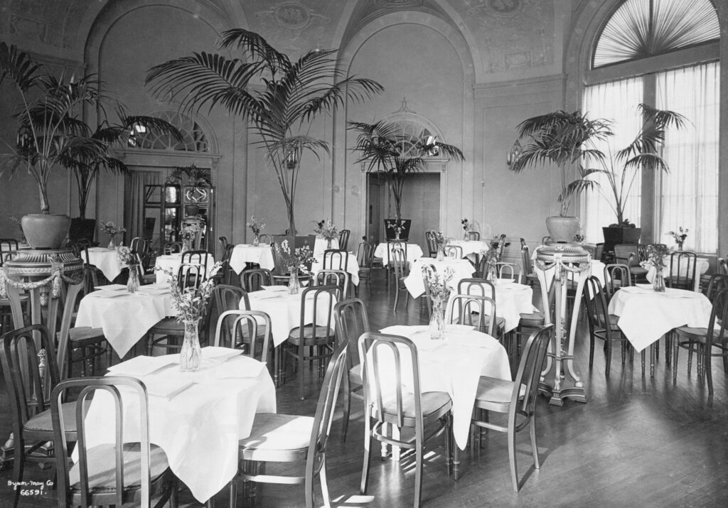 A black and white photograph of a circular banquet room in the Hotel MacDonald. The room is filled with round 4-person tables. Each table has a white tablecloth and a small bouquet in a clear glass vase. Surrounding the perimeter are palm trees in large pots.  