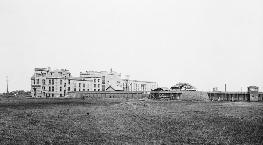 The three-storey cell block is pictured in a large field with surrounding fences and small outbuildings under construction.