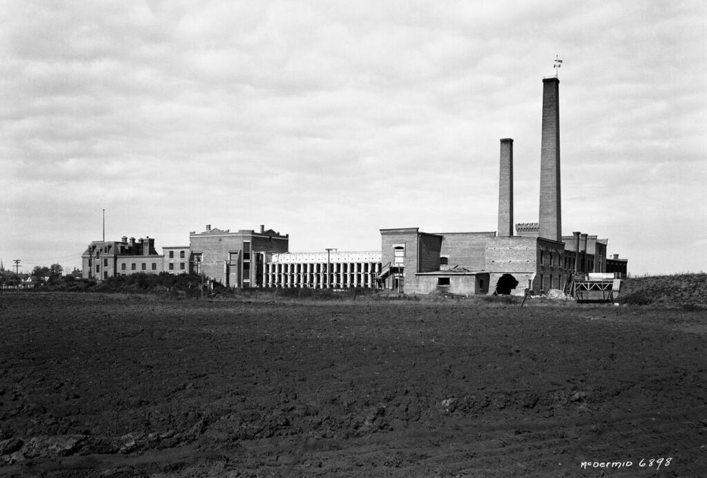 : A three-storey brick cell block is connected to a workshop and another large industrial building with two tall chimneys. There is a large hole in the side of one of the buildings suggesting it had started to be dismantled and/or suffered structural damage. A tilled farm field that would have been worked by prisoners is in the foreground.