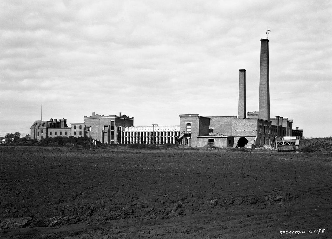 : A three-storey brick cell block is connected to a workshop and another large industrial building with two tall chimneys. There is a large hole in the side of one of the buildings suggesting it had started to be dismantled and/or suffered structural damage. A tilled farm field that would have been worked by prisoners is in the foreground.