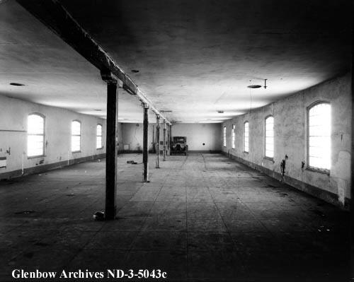 A long worn-down room with large windows on either side and five support pillars running down the middle of the room. There is a car parked at the end of the room with the front of the car facing the photographer.