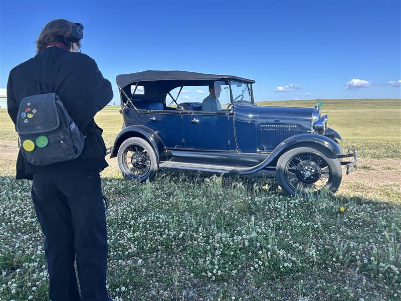 Emily stands with their back to the camera, headphones on, next to a restored Ford Model T with a man in the drivers’ seat.