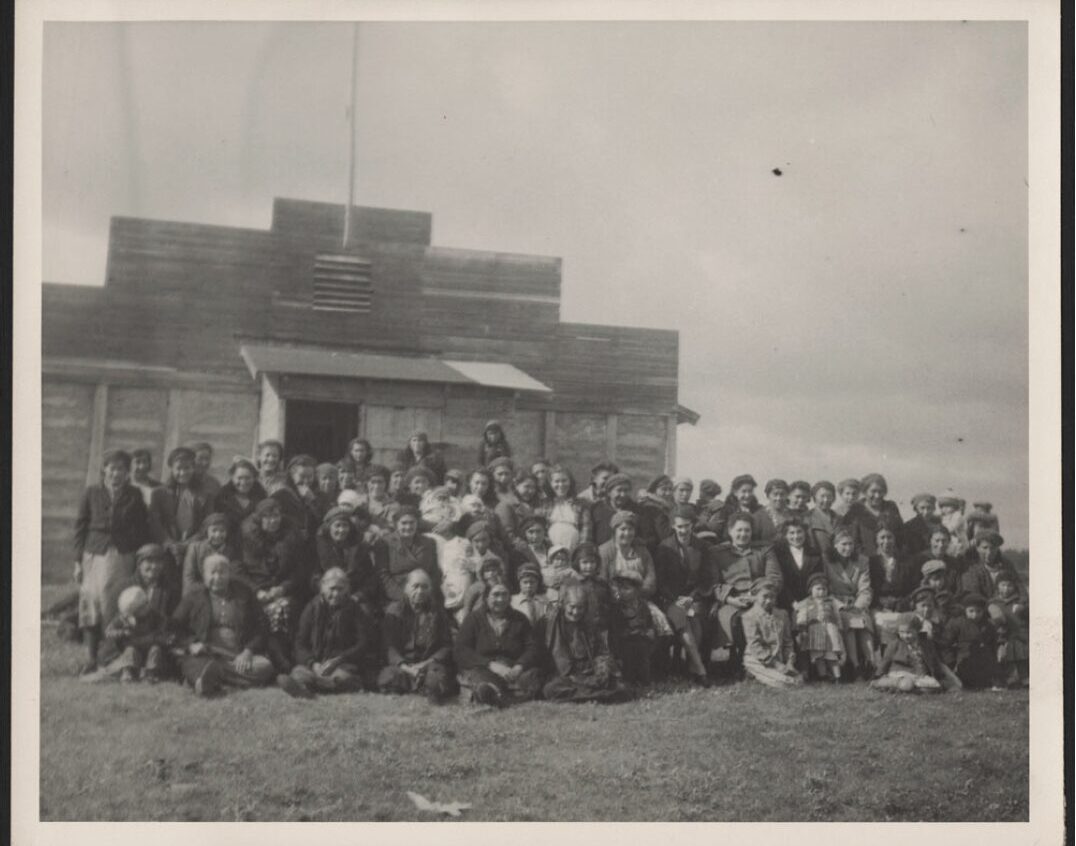 Around fifty women gathered in front of a wooden building. Included in the crowd are young children, smiling mothers, and older women. They are dressed in simple but comfortable-looking clothes, with some women holding babies.