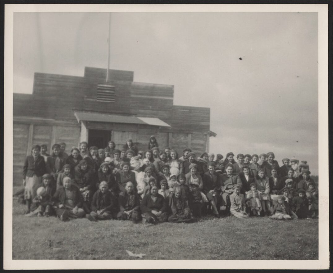 Around fifty women gathered in front of a wooden building. Included in the crowd are young children, smiling mothers, and older women. They are dressed in simple but comfortable-looking clothes, with some women holding babies.